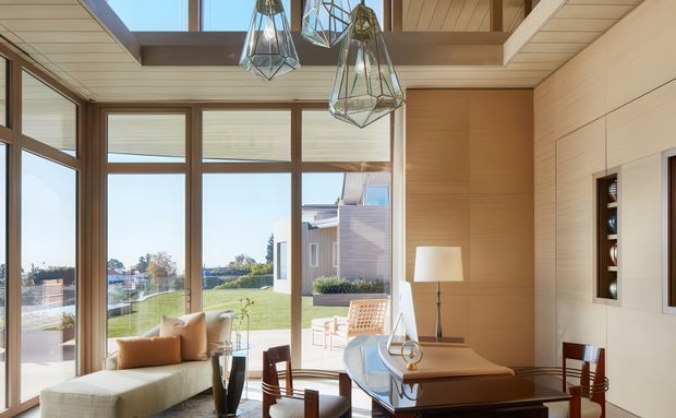 Bright, refined open-plan living and dining space with extensive floor-to-ceiling glazing overlooking a landscaped setting. The room features warm timber wall and ceiling finishes, a high ceiling with skylight-like glazing, sculptural geometric pendant lights, a curved upholstered window seat, a glass-topped dining table, and timber dining chairs. The architecture blends strong indoor-outdoor connection with natural materials and clean contemporary detailing.