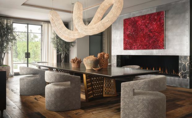 A sophisticated open-plan dining area with a large dark stone dining table, sculptural upholstered chairs, and an ornate textural chandelier. The room features warm timber flooring, full-height glazed doors with curtains, a dramatic red artwork, and a contemporary fireplace clad in dark metal or stone. The overall composition is luxurious, sculptural, and warmly atmospheric.