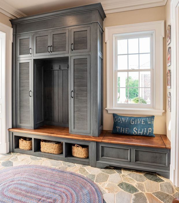 A bright, traditional entryway featuring a substantial dark-painted built-in mudroom unit with louvred cupboard doors, an open coat nook with hooks, a timber bench seat, woven storage baskets, and closed lower cabinetry. A multi-pane sash window sits above the bench, with warm beige walls, decorative ceiling cornices, and an irregular natural-stone floor. A round woven rug and nautical-themed cushion add casual character.