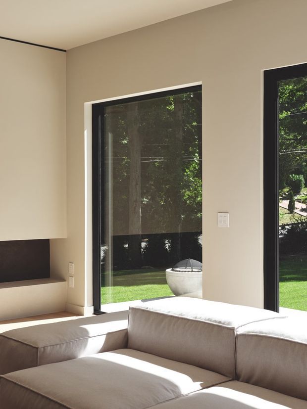 Bright, minimalist living room corner with a large low-profile sectional sofa in a neutral beige tone. Floor-to-ceiling black-framed glazing opens the room visually to a leafy garden, while a simple built-in wall niche or fireplace surround is visible at left. The room has smooth warm-neutral walls, a plain ceiling, strong natural daylight, and clean contemporary detailing.