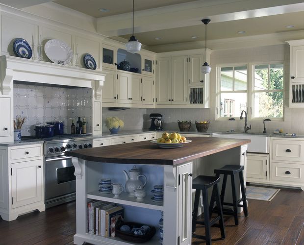 A bright, traditional-style kitchen with extensive cream-white shaker cabinetry, decorative plate display shelving, a tiled backsplash, stainless steel range, and dark timber flooring. A large central island has a dark timber benchtop, open display shelving, seating for three, and pendant lights above. The room includes a farmhouse-inspired sink area beneath a large window and detailed built-in joinery.