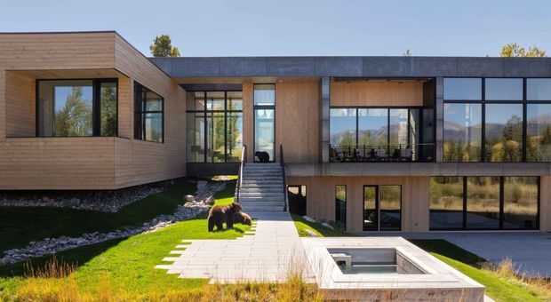 Contemporary two-storey residence with a low, flat-roofed composition, extensive dark-framed glazing, and warm horizontal timber cladding. The home steps around a landscaped courtyard with a central stair, paved terrace, and a sunken rectangular water feature or pool. Mountain and tree views are visible beyond the property.