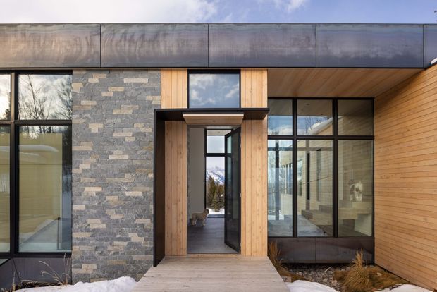 Exterior view of a contemporary mountain dwelling entrance, featuring dark metal roofing and window frames, natural vertical timber cladding, a stacked stone wall, extensive glazing, and a timber entry deck. Snow and a reflected mountainous landscape reinforce the alpine setting.
