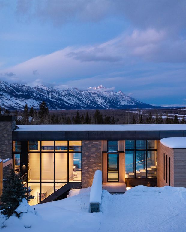 A contemporary flat-roofed residential building viewed from a snow-covered foreground at dusk, set against a dramatic snow-capped mountain range. The facade combines extensive dark-framed glazing, warm interior lighting, dark horizontal roof and wall elements, and sections of textured masonry and light timber-like cladding. The image strongly emphasizes the mountain setting and the building’s relationship to the snowy landscape.