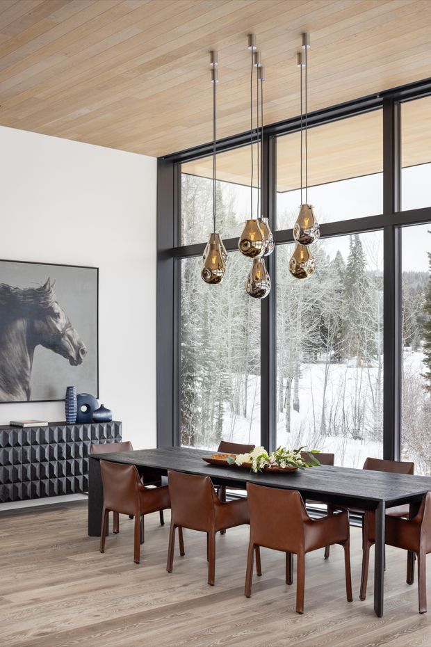 A refined dining area with a long dark rectangular dining table and multiple brown leather chairs, positioned beside expansive black-framed floor-to-ceiling windows overlooking a snowy wooded landscape. The room features a light timber-lined ceiling and matching timber flooring, a faceted dark sideboard with sculptural decor, a large horse artwork, and a cluster of suspended metallic pendant lights. The interior has a contemporary, warm, high-contrast aesthetic with strong indoor–outdoor connection.