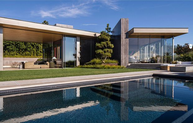 A modern single-storey residence viewed from a landscaped pool area. The home has strong horizontal flat roof planes, extensive floor-to-ceiling glazing, dark vertical wall elements, warm timber soffits, and a central textured stone or concrete feature wall. A reflecting swimming pool dominates the foreground, with outdoor seating visible through the glazing.