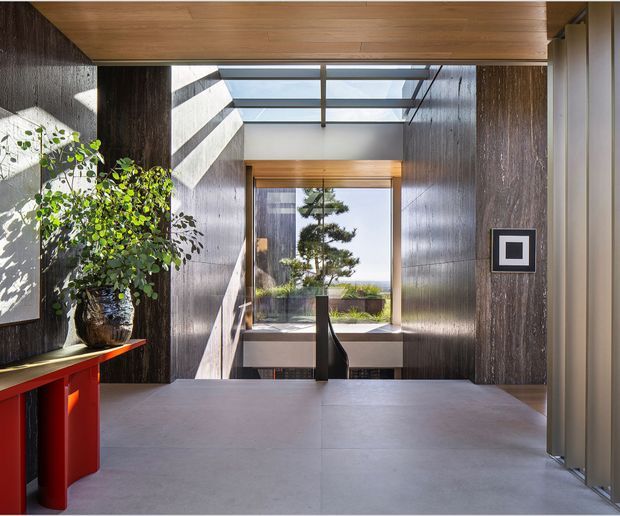 A refined contemporary entry hall with dark vertically textured wall panels, a broad grey floor, warm timber ceiling and door framing, and a central skylight that floods the space with daylight. A large glazed opening looks toward a landscaped exterior, while a vivid red console table and potted tree add contrast. Strong linear composition, deep shadow, and natural light create a dramatic yet welcoming arrival space.