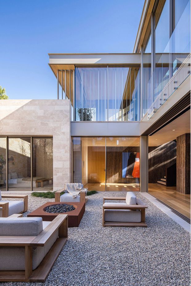 A modern multi-level residence organized around a gravel courtyard. The exterior combines pale stone walls, extensive floor-to-ceiling glazing, warm timber soffits and detailing, and dark vertical wood cladding. The courtyard contains low-profile timber-framed lounge seating and a central square fire pit, while a glazed entry and internal staircase are visible beyond.