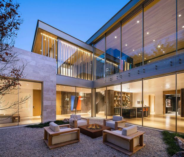 Twilight view of a sophisticated multi-level contemporary residence organized around a landscaped gravel courtyard. The home features extensive floor-to-ceiling glazing, warm timber-lined ceilings, pale stone-clad walls, slim dark frames, a sculptural vertical screen, and a sheltered outdoor seating area with a central fire feature. Warm interior lighting reveals living and dining areas through the glass.