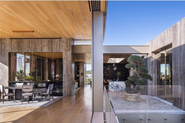 A refined contemporary residential interior centred on an open-plan dining area and a glazed courtyard. Warm timber lines the ceiling and flooring, while full-height glazing and dark textured wall surfaces frame a landscaped gravel courtyard with sculpted topiary. A long visual axis connects the dining room, hallway and outdoor courtyard, creating a bright indoor–outdoor composition.