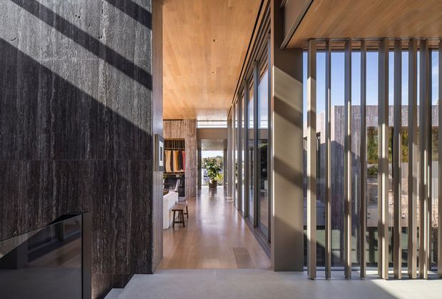 A refined modern residential interior showing a long entry hall or circulation space. Dark textured stone wall cladding and a built-in fireplace dominate the left side, while floor-to-ceiling glazing with vertical metal screening lines the right. Warm timber ceiling and timber flooring extend toward a bright living area visible at the end of the corridor. Strong sunlight creates graphic shadows across the stone and floor.