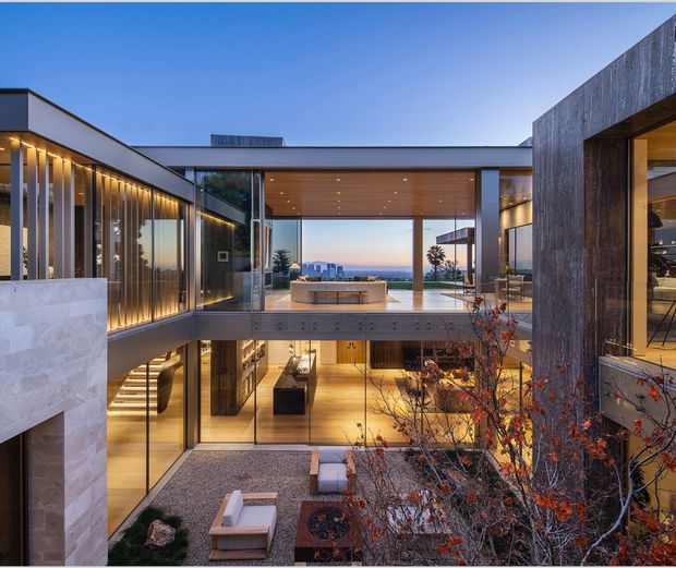 A contemporary multi-level residence organized around a landscaped courtyard, with extensive floor-to-ceiling glazing, projecting flat-roofed wings, and warm timber soffits. The view looks through the open central living area toward a distant urban and coastal horizon. Visible features include an upper-level outdoor terrace with built-in seating, a lower courtyard with gravel and lounge furniture, a glass bridge-like connecting volume, recessed lighting, and a mix of pale stone, dark textured cladding, timber, and concrete.