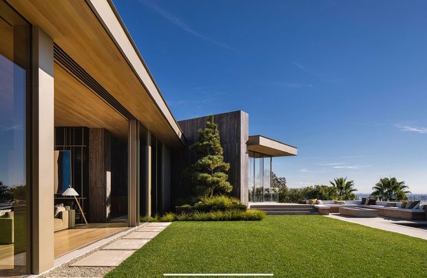Modern single-level residence viewed from a landscaped lawn, featuring expansive floor-to-ceiling glazing, warm timber soffits, dark vertical timber cladding, crisp rectilinear roof forms, and a stepped outdoor terrace with a pool or water feature overlooking a distant coastal or urban horizon.