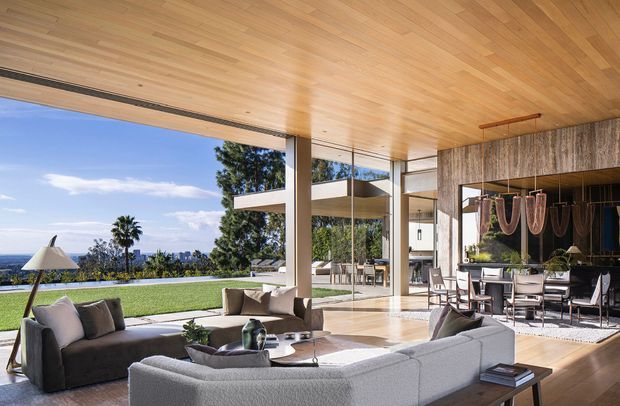 A luxurious open-plan living and dining space with extensive floor-to-ceiling glazing opening onto a landscaped lawn and distant coastal or urban views. The room features a warm timber-lined ceiling, pale stone or polished concrete flooring, neutral upholstered sofas, a dining table, and a dark textured feature wall with sculptural pendant lighting. Large structural columns and seamless indoor-outdoor connections create an airy, refined contemporary setting.