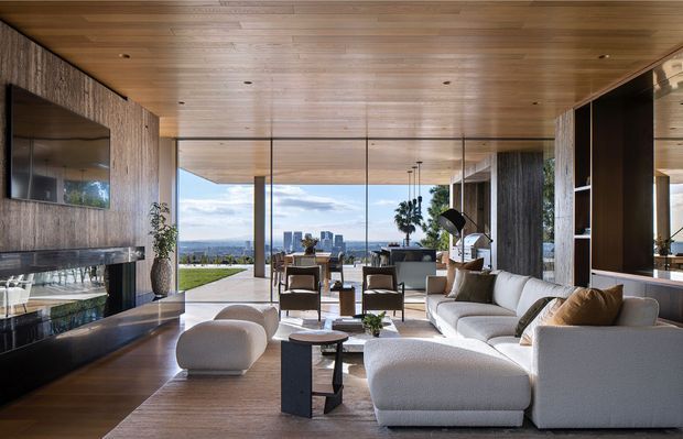 A sophisticated contemporary living room with a warm timber-lined ceiling, dark textured feature walls, extensive floor-to-ceiling glazing, and a panoramic urban view. The room includes a large light-toned sectional sofa, sculptural lounge chairs, minimalist coffee tables, a dark stone fireplace wall, integrated shelving, and a dining area beyond. The design has a refined indoor-outdoor connection and a balanced palette of timber, charcoal, cream, and grey.