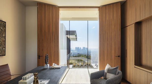 A refined home office with dark timber built-in cabinetry and tall paired timber doors opening to full-height glazing. A substantial desk faces a bright city and landscape outlook, with a lounge chair, desk accessories, and a small seating area in the foreground. The room has a warm, understated contemporary character and strong indoor-outdoor visual connection.