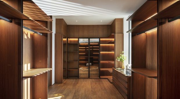 A sophisticated walk-in wardrobe or dressing room lined with dark timber built-in cabinetry, open shelving, drawers, and warm integrated LED lighting. A central glazed wardrobe display anchors the rear wall, while a narrow island or drawer unit sits to the right. Timber flooring and rich brown joinery create a warm, luxurious feel, with daylight filtered through blinds on the left.