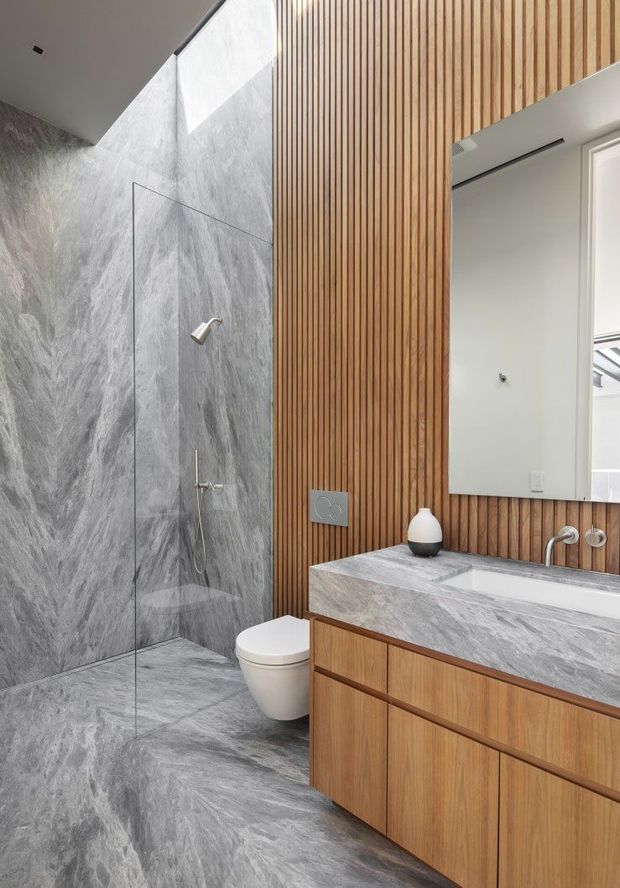 A contemporary bathroom featuring a walk-in shower enclosed by frameless glass, grey marble-look wall and floor surfaces, a wall-hung toilet, and a timber floating vanity with integrated grey stone-look benchtop and basin. Vertical timber battens form a warm feature wall behind the vanity and mirror. A skylight or high window provides natural light.