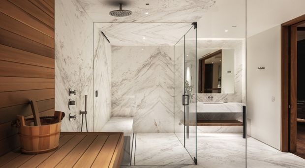 Luxurious contemporary bathroom featuring a glass-enclosed marble shower, a timber sauna bench and wall cladding in the foreground, and a floating timber vanity with a large mirror. The space uses veined white stone surfaces, warm wood, frameless glazing, and recessed lighting.
