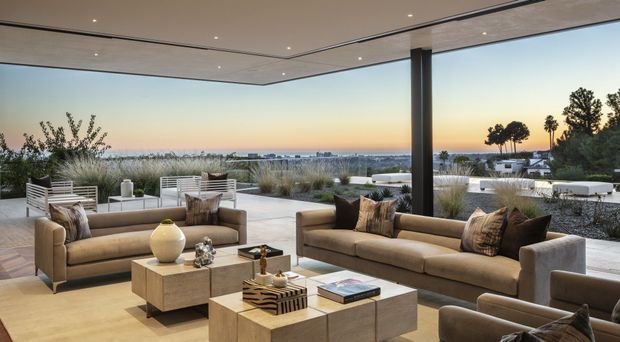 A refined, open-air living room terrace with expansive glazing-free views toward a coastal or urban horizon at sunset. The space features neutral upholstered sofas and armchairs arranged around low square coffee tables, a pale stone or concrete floor, a broad flat ceiling with recessed downlights, and a dark structural column. Built-in outdoor seating, ornamental grasses, and low landscaping frame the view. The overall setting is luxurious, calm, and contemporary.