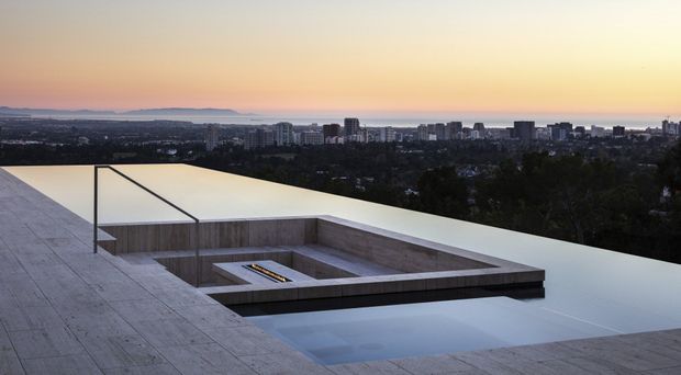 A minimalist rooftop terrace at dusk, featuring a large shallow reflecting pool or water feature integrated into a broad pale stone or concrete roof deck. Low rectangular parapets and a recessed stepped courtyard-like composition frame the pool, while slender dark metal posts and lines create a restrained architectural detail. A distant urban skyline and low hills are visible beyond the treed foreground.