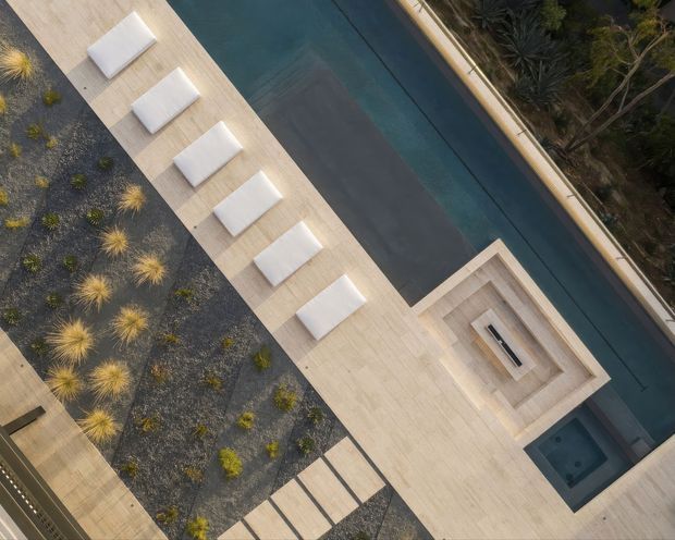 Aerial view of a contemporary outdoor pool area with a long rectangular swimming pool, pale timber-toned decking, a row of white sun loungers, a recessed square fire or seating feature, and dark gravel landscaping with ornamental grasses.
