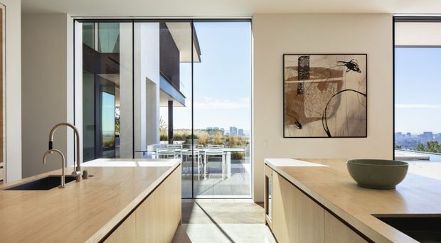 Bright contemporary kitchen with two large light-wood islands featuring pale stone benchtops, integrated dark sinks and sculptural brushed-metal tapware. Floor-to-ceiling glazing opens to a balcony or terrace and provides expansive urban views. Minimal cream walls, dark-framed glazing, and abstract wall art create a clean, refined interior.