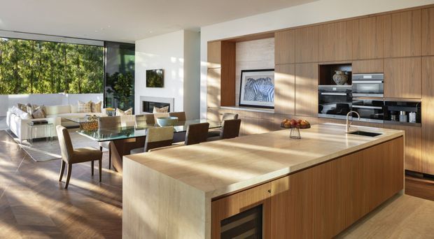Open-plan contemporary kitchen and dining area with a large timber island, integrated stainless-steel appliances, and extensive warm wood cabinetry. A glass dining table and living area sit beyond the kitchen, with floor-to-ceiling glazing overlooking greenery and bringing in abundant natural light. The space has clean lines, a warm neutral palette, and a refined indoor-outdoor feel.