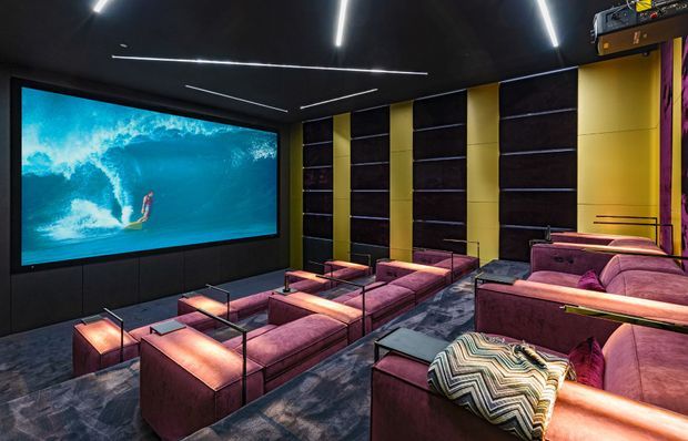 A dark, luxurious home cinema with tiered rows of plush burgundy and mauve reclining seating, a large wall-mounted screen displaying a surfing scene, dark acoustic wall panels with vertical mustard-yellow accents, black ceiling, linear strip lighting, and patterned carpet. The room has a dramatic, immersive entertainment-focused atmosphere.