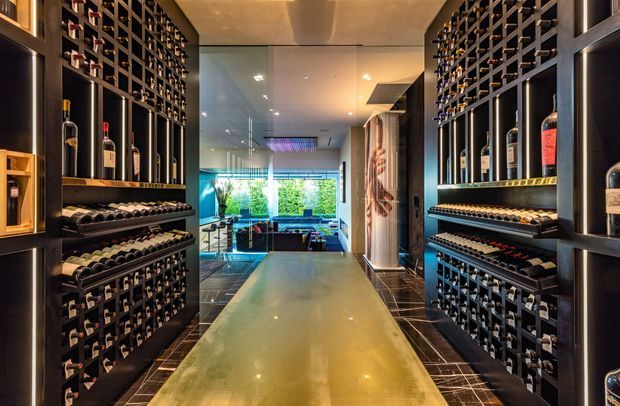 A narrow, sophisticated wine storage room or passage lined on both sides with dark built-in wine racks and illuminated bottle display shelving. A polished reflective central floor or walkway leads toward a glazed living or entertaining area at the rear, with warm recessed lighting, dark tiled flooring, and a contemporary luxurious atmosphere.