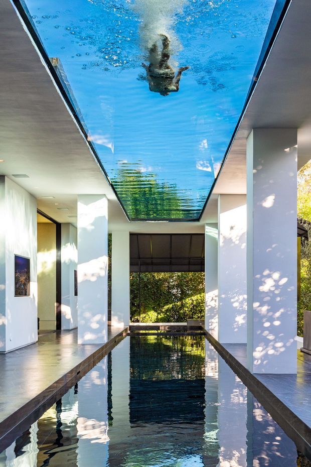 A dramatic contemporary interior featuring a long narrow reflecting pool or swimming pool running through a covered corridor. White structural columns and walls frame the water, with a large transparent overhead skylight or glass pool panel showing a swimmer above. Dark polished edging contrasts with the bright pool and sun-dappled surfaces, while greenery is visible beyond the open end.