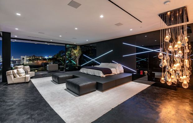 A dramatic, spacious contemporary bedroom with a low platform bed, dark upholstered ottomans, and a large pale area rug over dark flooring. Glossy black wall panels are accented by angular linear LED lighting, while a sculptural cluster chandelier with numerous glowing glass pendants hangs from the ceiling. Full-height glazing opens to a nighttime city view, with a small seating area beside it.