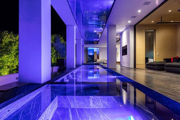 Dramatic nighttime view of a luxurious contemporary residence, looking along a long reflective indoor-outdoor water feature or pool beside an open-plan living area. Large white structural columns and floor-to-ceiling glazing frame landscaped greenery, while blue-violet lighting reflects across the water and polished stone surfaces. The living space contains a low dark sectional sofa, wall-mounted television and timber-toned flooring.