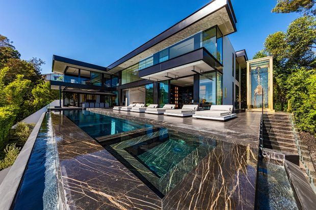 A dramatic contemporary multi-level residence set among mature trees, featuring extensive floor-to-ceiling glazing, cantilevered upper volumes, dark marble-clad terraces, and a reflective infinity-edge swimming pool. The angular architecture and strong black, charcoal, and glass palette create a luxurious, high-contrast exterior composition.
