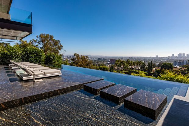 Luxury hillside outdoor terrace with a striking infinity pool overlooking a sprawling city skyline. The space features dark veined stone paving and stepped platforms, low dark stone tables or plinths, and a row of light-coloured sun loungers beside the pool. Part of a contemporary home with projecting roof and glazing is visible at the upper left, while landscaped planting frames the distant urban view.