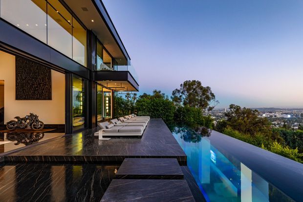 Luxury multi-level modern residence viewed from an elevated outdoor terrace, featuring extensive floor-to-ceiling glazing, dark stone paving and cladding, projecting balconies, an infinity-edge swimming pool, and a panoramic urban landscape beyond. The space is styled with streamlined outdoor loungers and dramatic evening lighting.