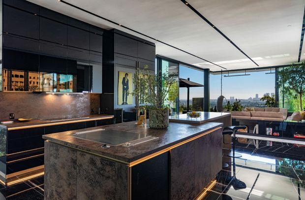 A sophisticated open-plan kitchen with a large central island, dark built-in cabinetry, and a dramatic stone-look island and splashback. Warm integrated LED strip lighting highlights the cabinetry and island edges. Floor-to-ceiling glazing opens toward an outdoor terrace and distant urban view, while a dining or lounge area is visible beyond. The space has a luxurious, contemporary feel with strong black, charcoal, bronze, and warm timber-toned accents.