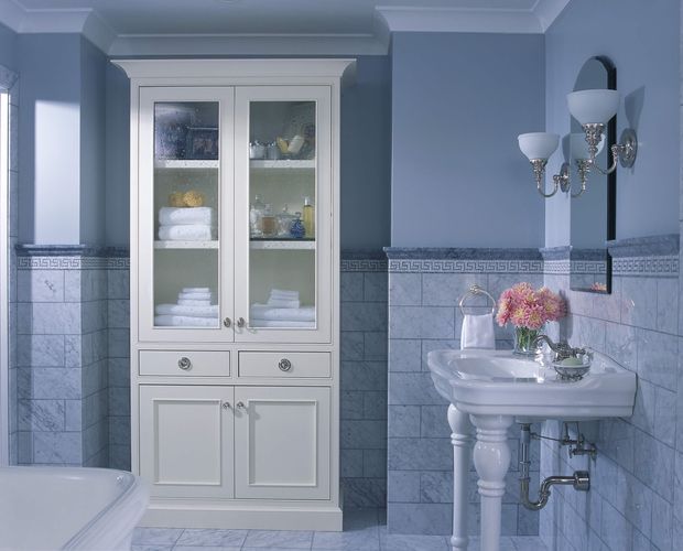 A traditional bathroom in a cool blue and white palette, featuring a pedestal sink, wall-mounted mirror, crystal-style wall sconces, tiled walls with a Greek key border, a freestanding storage cabinet with glass doors, and the edge of a bathtub. Decorative mouldings and classical plumbing details give the room a refined, period-inspired character.