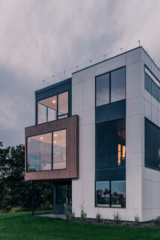 Exterior view of a contemporary multi-storey residence with a crisp white rectilinear facade, dark-framed floor-to-ceiling glazing, and a projecting timber-clad balcony or bay volume. The building sits beside a landscaped lawn and trees under an overcast sky, with warm interior lighting visible through the windows.