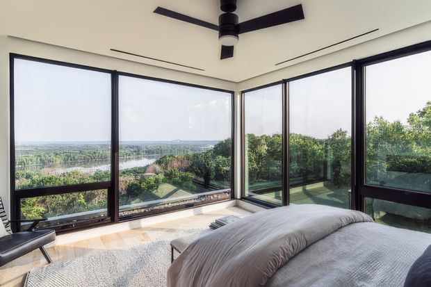 Bright contemporary bedroom with a low-profile bed facing expansive corner glazing. Dark-framed floor-to-ceiling windows wrap around the room, opening to a landscaped garden and distant horizon. The space has pale walls, light timber flooring, a large neutral rug, minimal furnishings, and a black ceiling fan.
