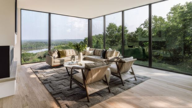 Bright contemporary living room with a generous seating arrangement centred on a coffee table and rug, enclosed by floor-to-ceiling corner glazing. Pale timber flooring, neutral upholstered sofas and armchairs, and slim dark window frames create an airy interior with expansive views toward a wooded landscape and water beyond.