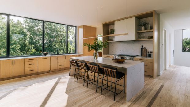 A bright, open-plan contemporary kitchen with extensive light timber cabinetry, a large pale stone island with seating for four, integrated appliances, and floor-to-ceiling black-framed glazing overlooking leafy greenery. Light timber flooring, white walls, open shelving, and indoor plants create a warm, airy atmosphere. A hallway and adjacent room are visible beyond the kitchen.