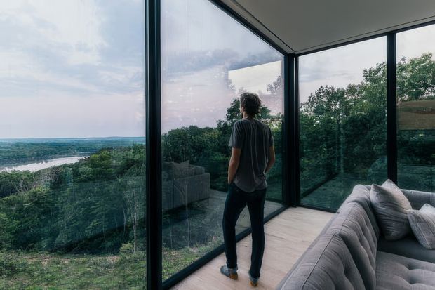 A modern interior corner with full-height, dark-framed glazing wrapping around a panoramic view of dense greenery, a river, and distant landscape. A person stands near the windows beside a large curved grey sectional sofa on a light timber floor. The space feels contemporary, calm, and strongly connected to the surrounding landscape.