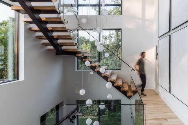 A bright, contemporary stairway with floating timber treads supported by a dark steel structure. The open stair descends through a tall multi-level void, enclosed by clear glass balustrades and surrounded by large windows. Suspended spherical pendant lights hang through the stairwell, while white walls and light timber flooring create an airy, minimalist interior.