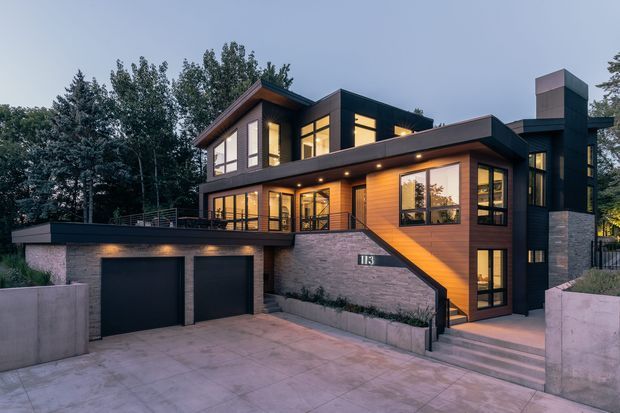 Contemporary multi-level residence photographed from the front at dusk. The house features dark horizontal cladding, warm timber-toned wall panels, extensive black-framed glazing, projecting flat roof forms, a stone-clad lower level, and an integrated double garage. Broad concrete paving, retaining walls, stairs, and planted edges define the approach, while warm interior lighting glows through the windows.