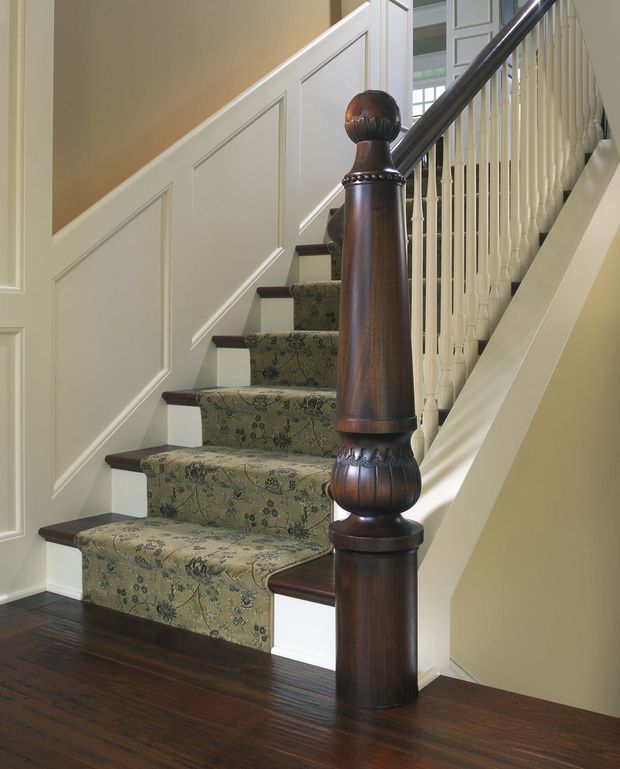 A close interior view of a traditional staircase with an ornate dark timber newel post and handrail, white turned balusters, and patterned green stair runner. The staircase has white painted panelled wainscoting and trim, dark timber stair treads, and dark timber flooring, creating a formal, heritage-inspired entry or circulation space.