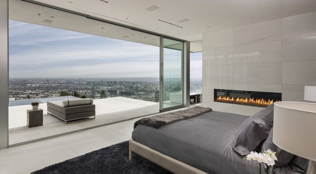 A luxurious contemporary bedroom with a large bed facing floor-to-ceiling corner glazing and an expansive city view. The room has a minimalist white interior, pale flooring, a long linear built-in fireplace, a freestanding bathtub partially visible at right, and a terrace with outdoor seating beyond the glass.