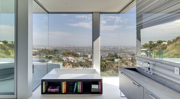 Contemporary open-plan kitchen and bathroom-like bathing area with expansive floor-to-ceiling glazing overlooking a hilly urban landscape. A freestanding white rectangular bath sits centrally on a dark built-in plinth with open shelving, while a linear kitchen bench with integrated cabinetry, sink and tap occupies the right side. The space has a flat ceiling with recessed downlights, pale grey surfaces and strong indoor-outdoor visual connection.