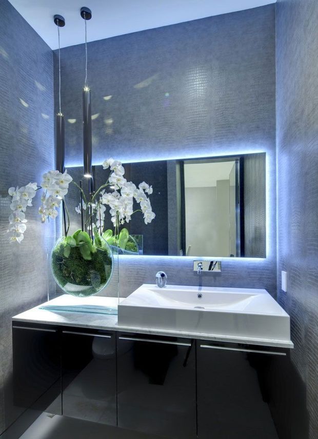 Compact contemporary bathroom with a floating glossy black vanity, white integrated basin, large backlit mirror, dark textured wall finishes, and sculptural pendant lighting. A glass shower screen and orchid arrangement add decorative detail, while cool LED illumination creates a polished, luxurious atmosphere.