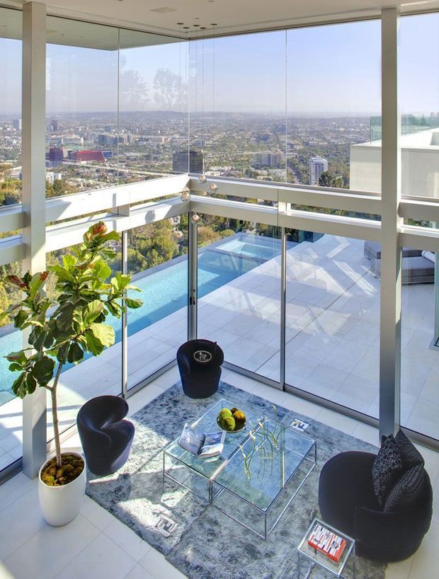 A bright, double-height glazed living area with floor-to-ceiling windows overlooking a dense cityscape. The room has pale tiled flooring, white structural columns and framing, a glass coffee table on a grey rug, sculptural black lounge chairs, and a potted indoor tree. An outdoor terrace and swimming pool are visible through the glazing below.